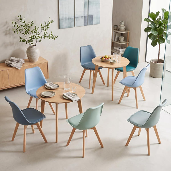 A set of modern dining chairs and round wooden tables arranged in a bright, minimalist dining room setting. The chairs feature molded plastic seats in shades of light blue, teal, and mint green with light natural wood legs.