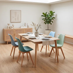 A modern dining room scene with a rectangular light wood table surrounded by four chairs in teal, mint green, and light blue, set on a light wood floor against cream walls.