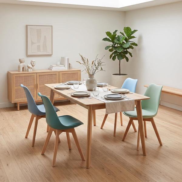 A modern dining room scene with a rectangular light wood table surrounded by four chairs in teal, mint green, and light blue, set on a light wood floor against cream walls.