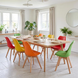 A modern dining room scene with a light wood rectangular table surrounded by colorful molded chairs in red, yellow, orange, and green, set with tableware and surrounded by potted plants.