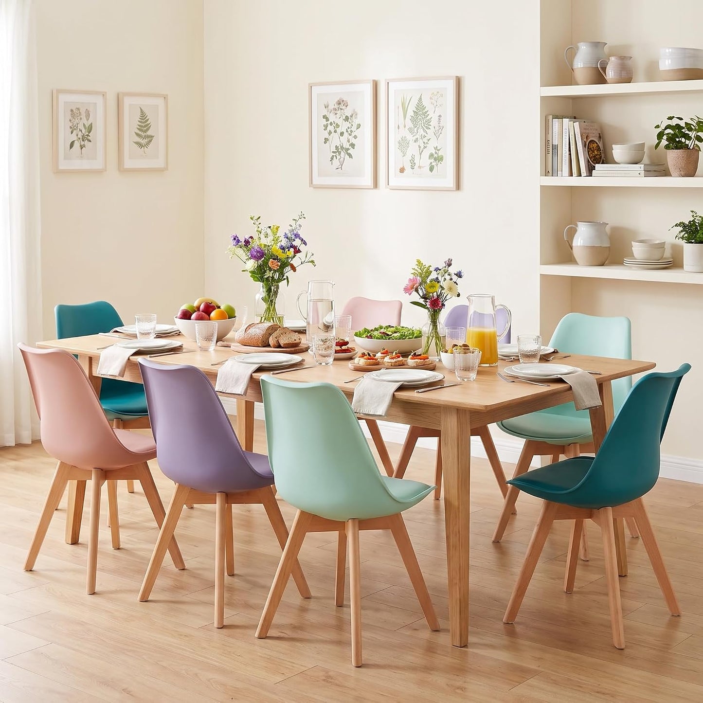 A dining room scene with a rectangular light wood table surrounded by eight pastel-colored chairs in pink, blue, purple, mint green, and teal. The table is fully set with plates, cutlery, glasses, and floral centerpieces, set against a warm cream wall with botanical prints and built-in white shelves.