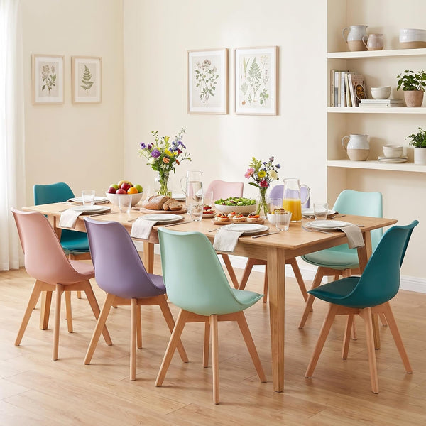 A dining room scene with a rectangular light wood table surrounded by eight pastel-colored chairs in pink, blue, purple, mint green, and teal. The table is fully set with plates, cutlery, glasses, and floral centerpieces, set against a warm cream wall with botanical prints and built-in white shelves.